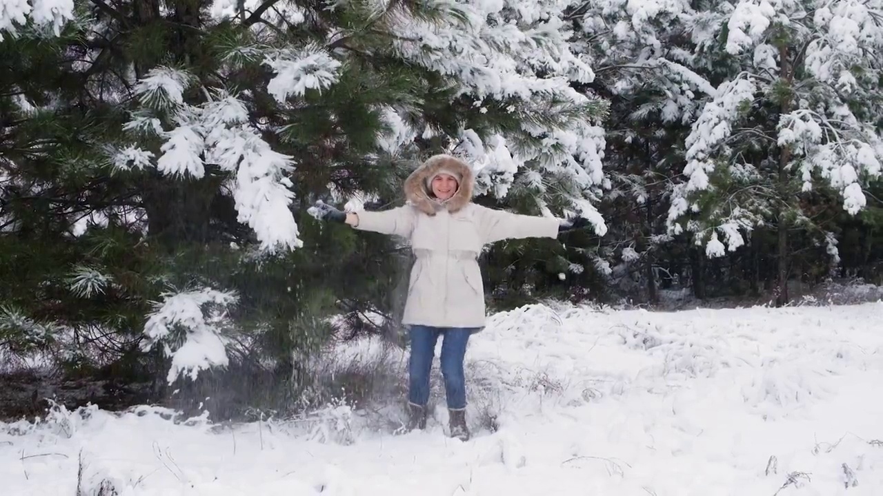 在冬天的松树林中，一名穿着白色夹克的女子在雪下的树枝下跳跃嬉戏视频素材