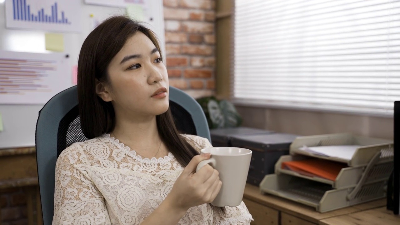 疲惫的亚洲女商人凝视着远方，闭上眼睛，坐在办公室的椅子上喝着咖啡休息。视频素材