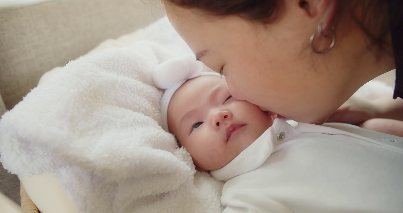 幸福家庭年轻的亚洲母亲抱着她漂亮的新生婴儿在家里的床上亲吻着婴儿，孩子，父母，童年，生活，母性，母爱的概念视频素材