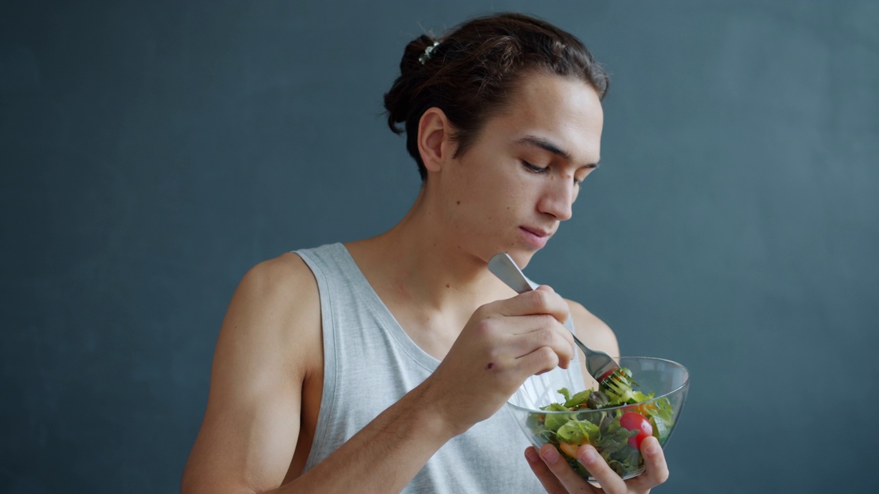 慢动作的肖像年轻的千禧一代吃蔬菜沙拉享受健康的食物在灰色的背景视频素材