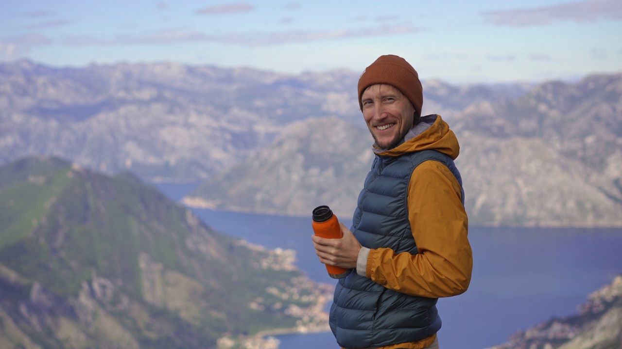 一名男子秋天在黑山旅行。他一边喝着热水瓶里的热饮，一边站在俯瞰Boka Kotorska湾的一个观景点上视频素材