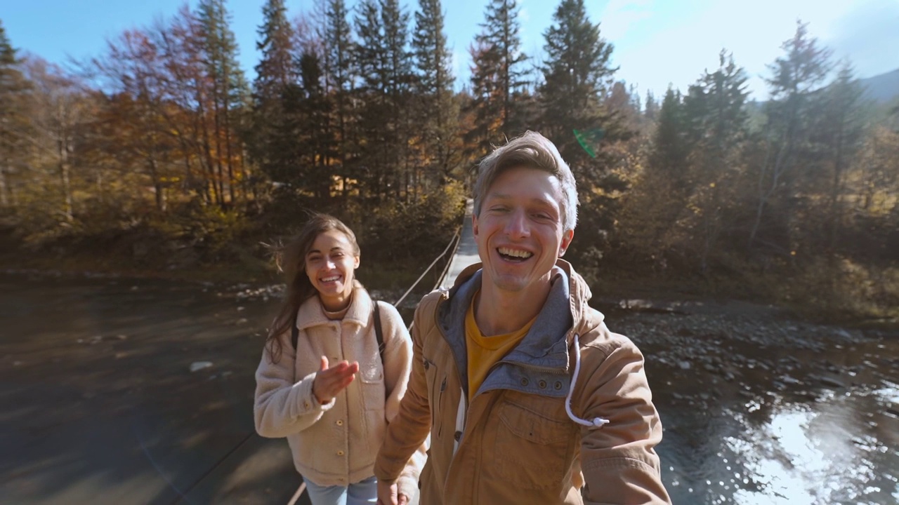 一对快乐的徒步旅行者在悬挂在山河上的木制吊桥上散步。男人和女人在令人惊叹的风景上自拍，背景是云杉、山脉和天空视频素材