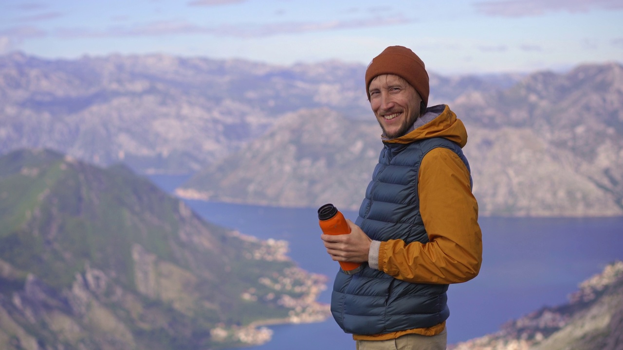 一名男子秋天在黑山旅行。他一边喝着热水瓶里的热饮，一边站在俯瞰Boka Kotorska湾的一个观景点上视频素材