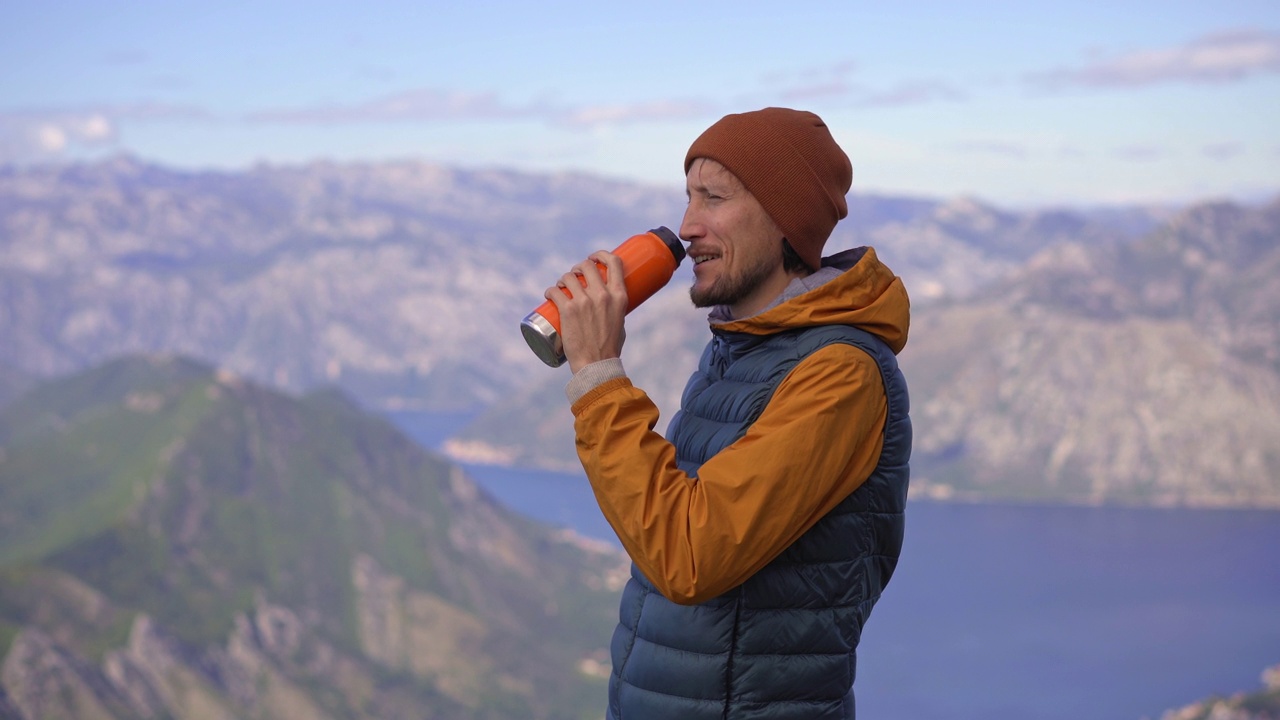 一名男子秋天在黑山旅行。他一边喝着热水瓶里的热饮，一边站在俯瞰Boka Kotorska湾的一个观景点上视频素材