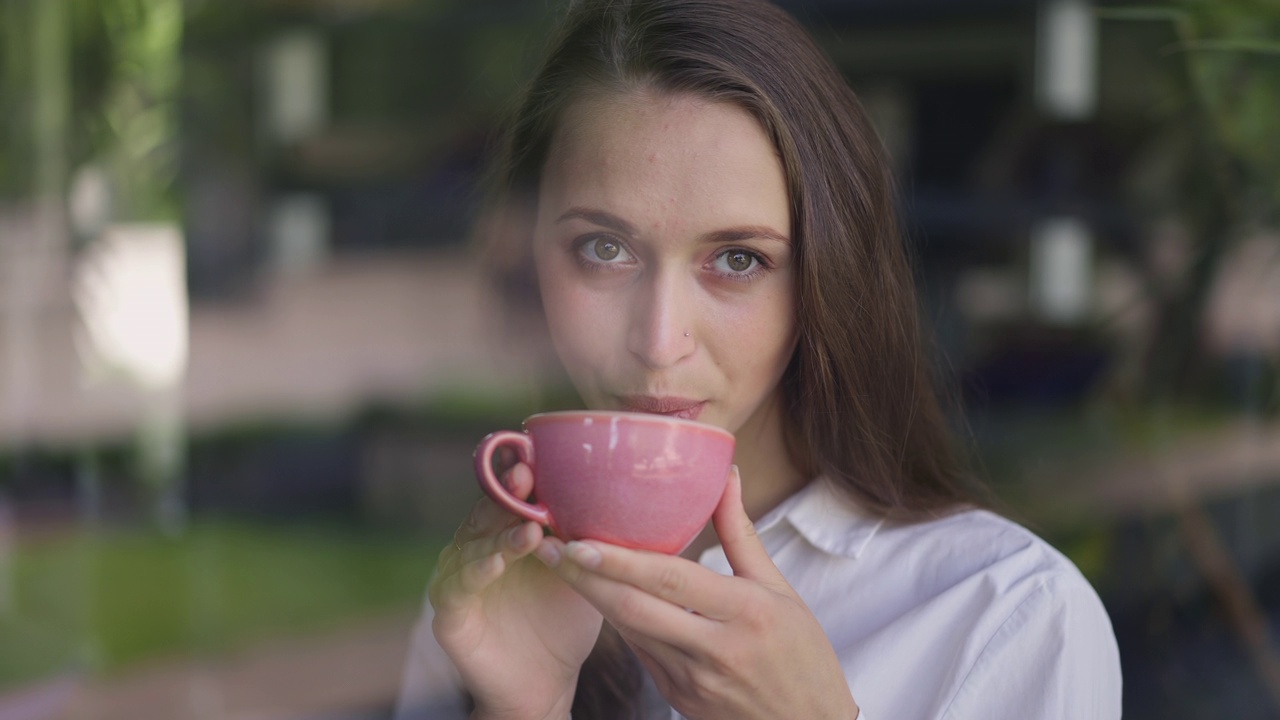 深色皮肤的白人妇女与棕色的眼睛看着窗外喝咖啡在咖啡馆的慢动作。城市里悠闲放松的美女周末喝热饮的写真视频素材