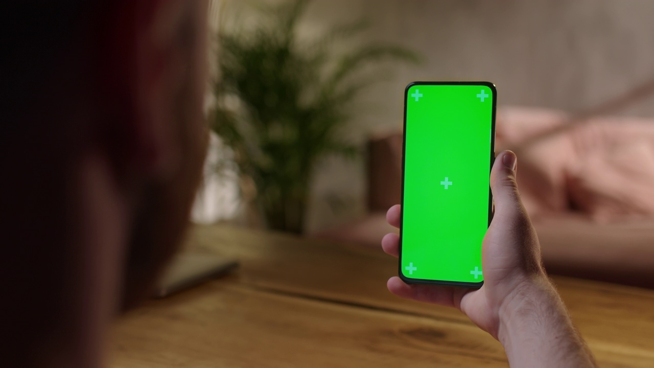 手持相机:后视图的年轻人坐在家里的一个木桌子上使用绿色模拟屏幕智能手机。男孩在不看电子设备屏幕的情况下观看内容视频素材