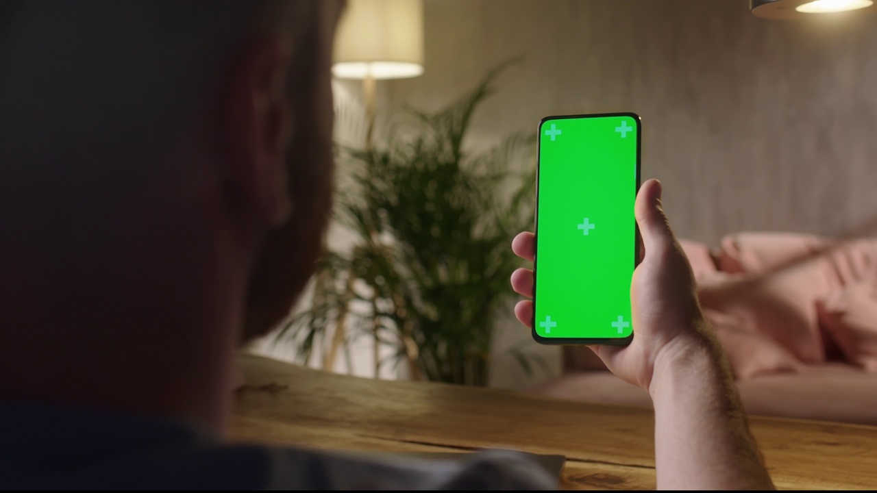 手持相机:在现代房间的人坐在使用电话与绿色模型屏幕色度键跟踪点上网观看内容视频博客的后视图。刷卡了视频素材