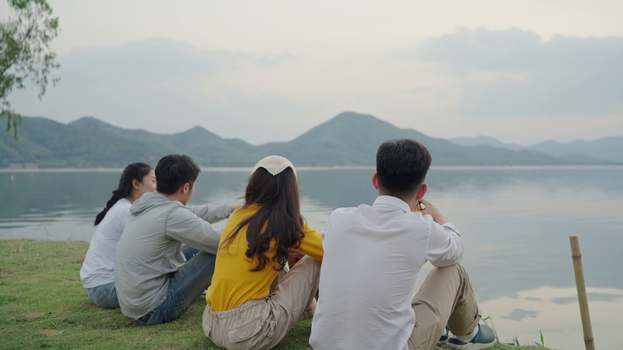 后视图。群亚洲四青年朋友男女在河边野餐，他们聊天，笑着，喝着庆祝欢呼啤酒视频素材