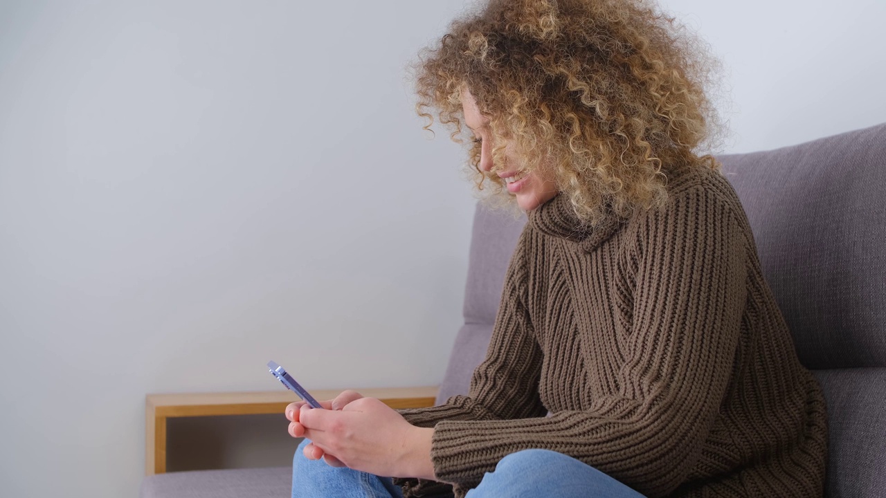 白人女人在用电话。一头卷发的年轻成年女性在封锁期间坐在家里的沙发上，在智能手机上的社交媒体应用程序上打字。房间里有现代手机的人视频素材