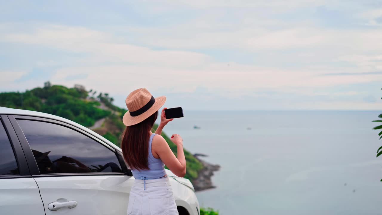 年轻的女性旅行者看着和拍照在美丽的海景与她的车，而旅行驾驶公路旅行的假期视频素材