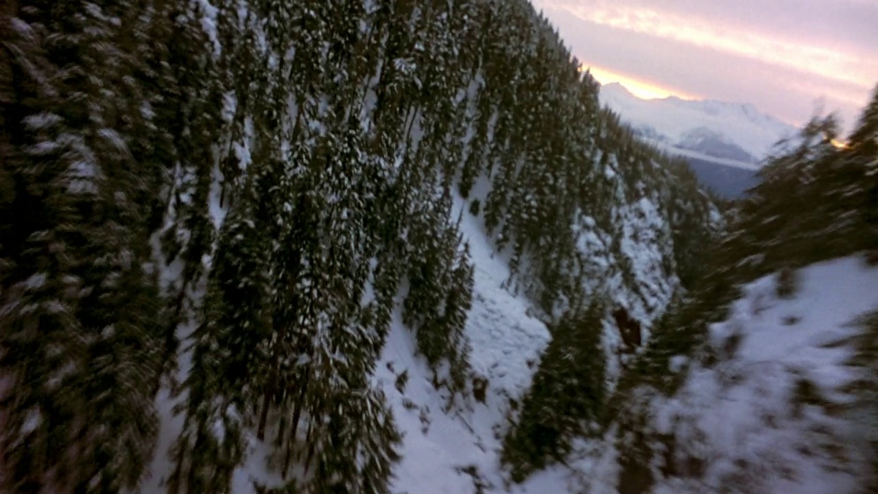 空中穿越雪山。镜头从喷气式飞机快速穿越山脉的角度旋转而来。看那绿油油的松树，上面撒满了雪。天空是厚厚的白色的雾和云。飞机在山区蜿蜒穿过山谷视频素材