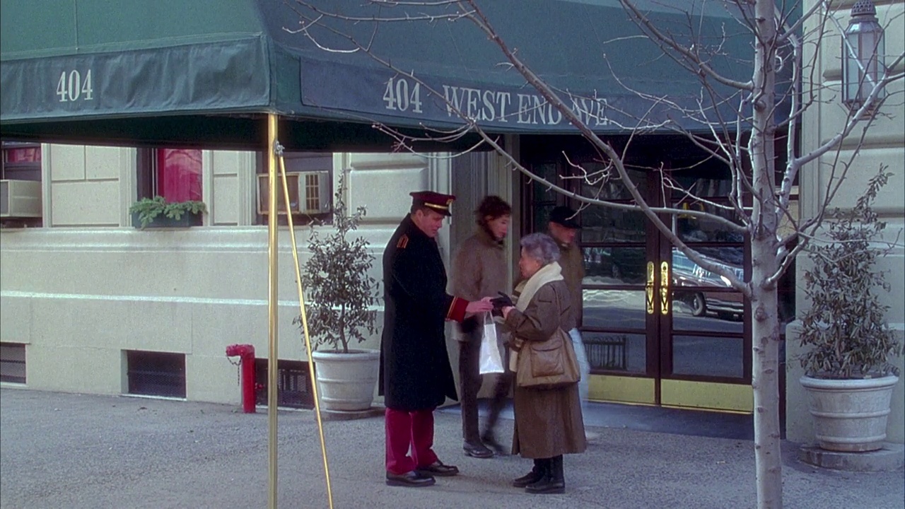 公寓大楼入口和雨篷的中角，雨篷上写着“404 west end ave.”老妇人向看门人展示了一个购物袋和一个盒子，他们一起走进了门。摇到三楼的窗户上。视频素材