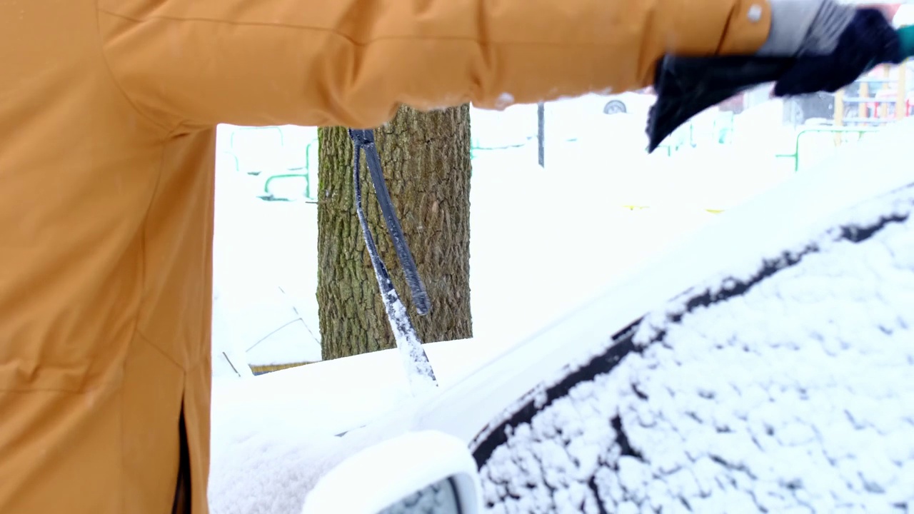 一名男子在雪后从车上刷雪。一只穿着芥末色夹克的手，白色的身体上放着汽车扫把。冬天的天气条件。慢动作视频素材