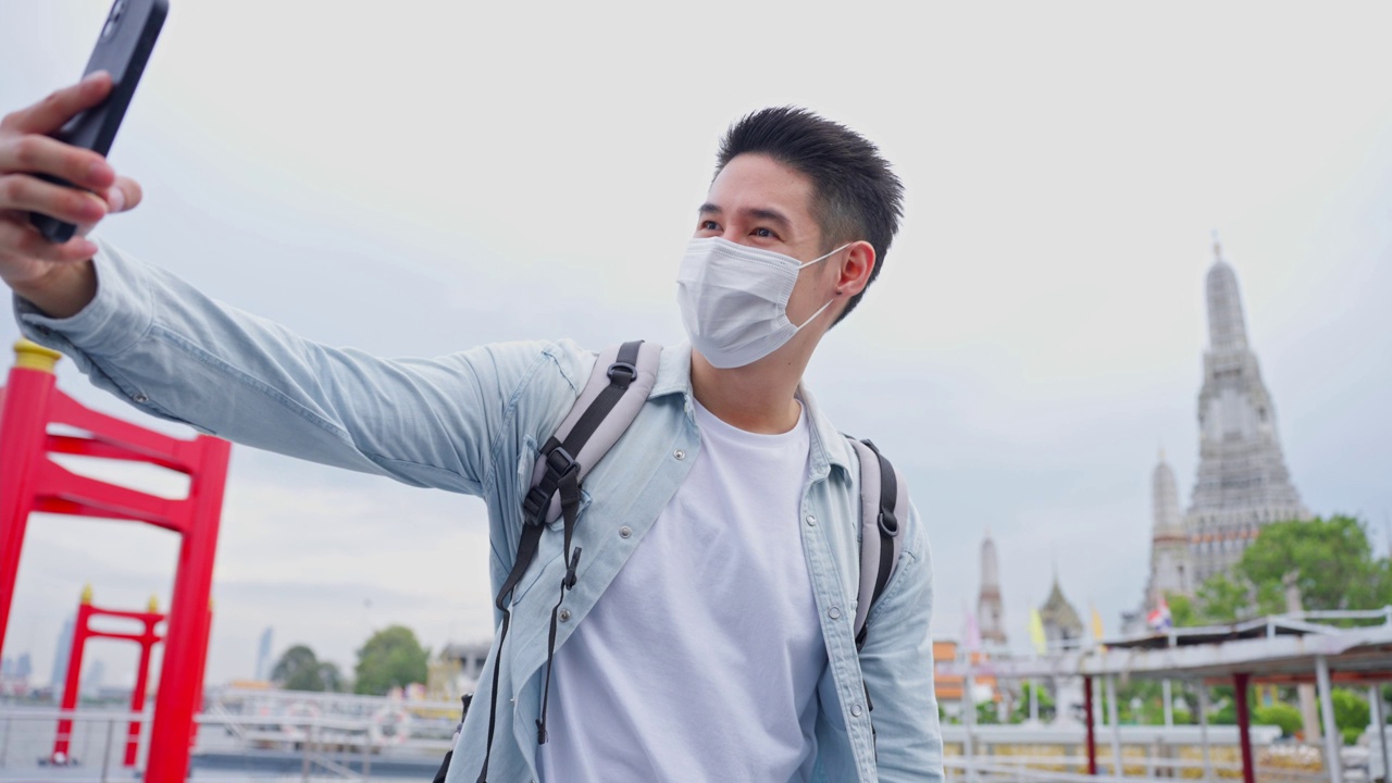 亚洲年轻男性背包客在城市里用智能手机自拍。帅哥旅游旅行者戴着口罩独自在街上旅行，用智能手机录视频，在泰国度假旅行视频素材