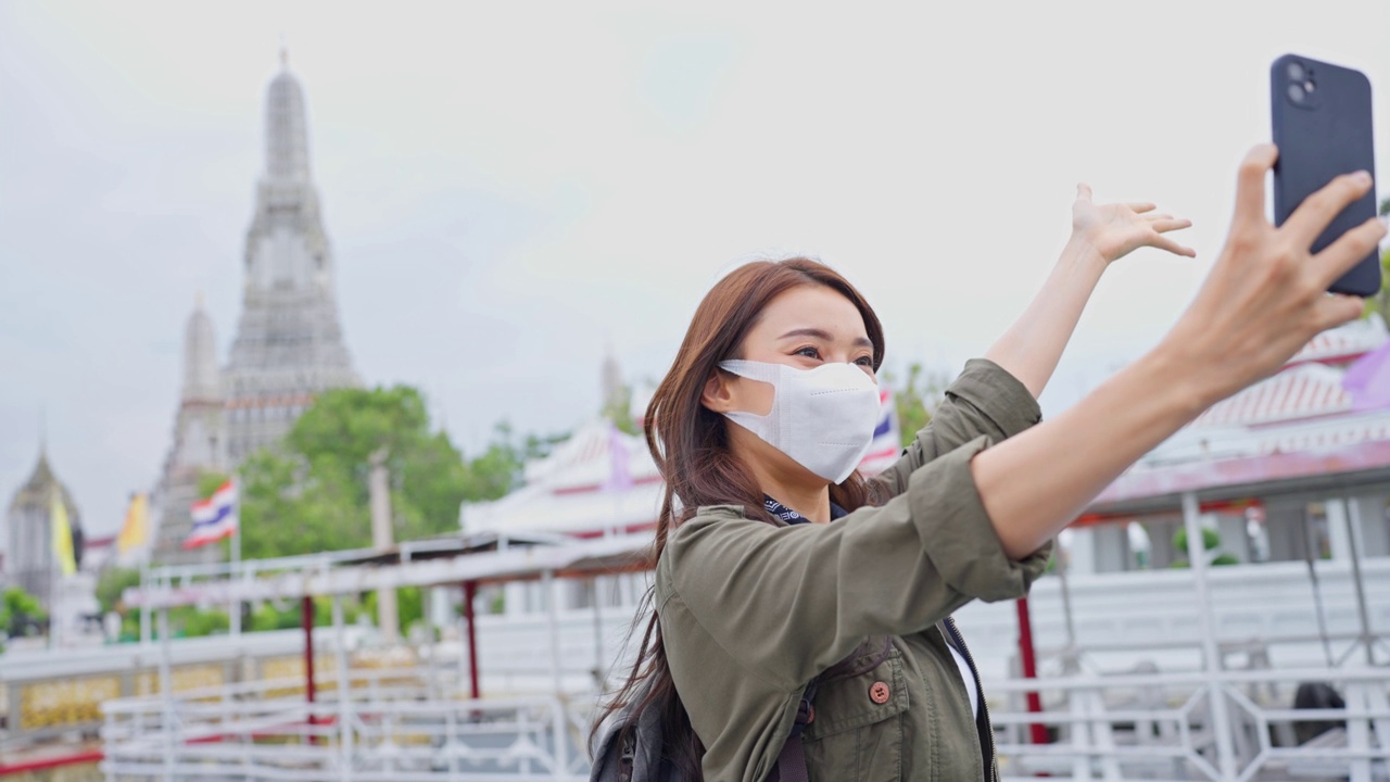 亚洲女性背包客用手机自拍，在城市里拍照。美丽的女游客戴着口罩独自在街上旅行，在泰国度假时用智能手机录视频视频素材