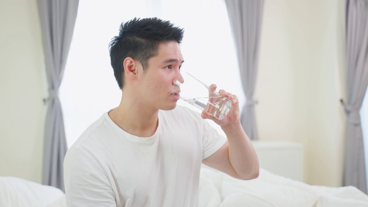 亚洲活跃的强壮男人的肖像在卧室里拿着干净的水。吸引人的口渴男性饮料或喝一小口在杯子的天然矿物质，为了健康和幸福醒来后看着相机。视频素材