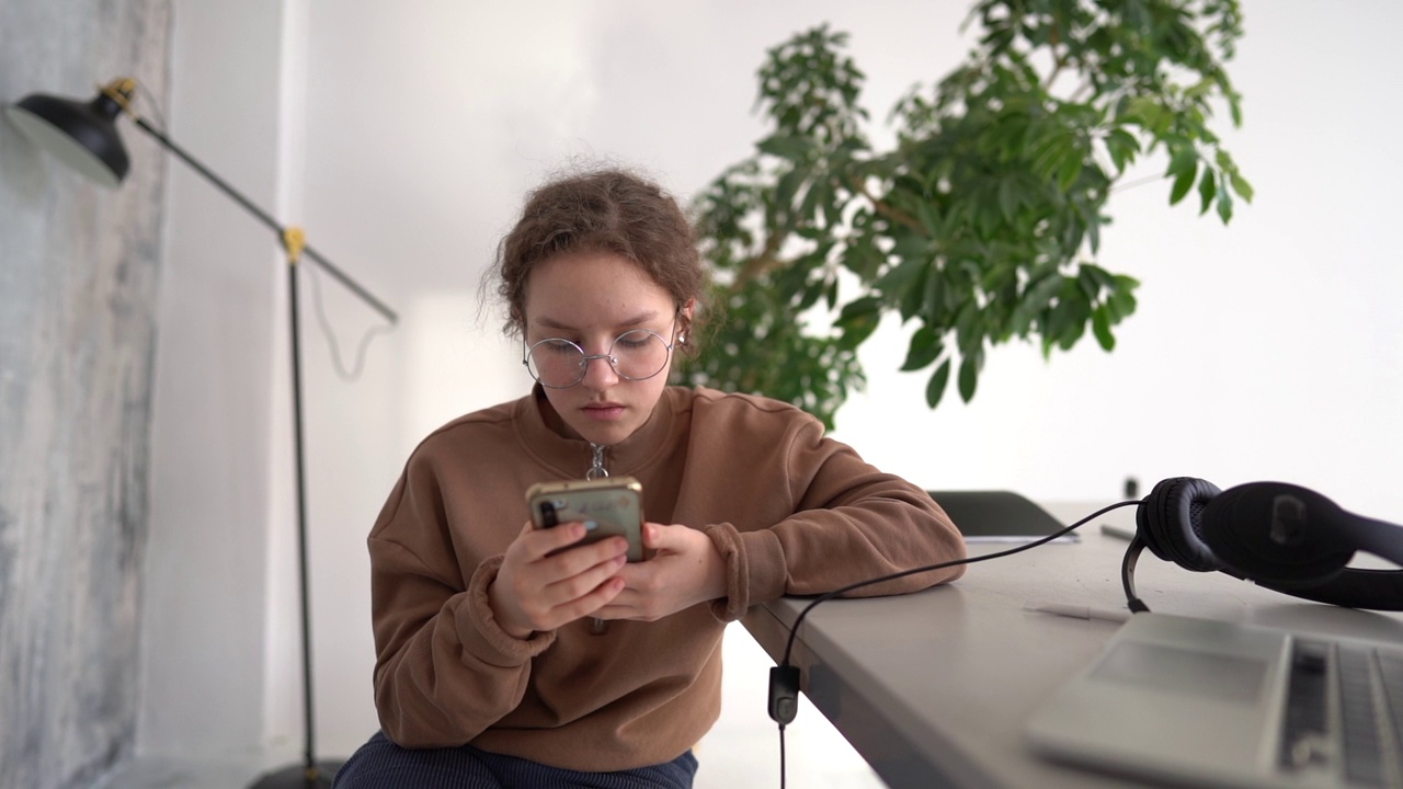 一个十几岁的女孩戴着眼镜用她的智能手机的特写。这个女孩在家里与朋友交流。封锁期间的远程学习视频素材