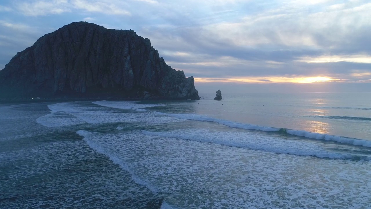 镜头从海滩开始，穿过汹涌的海浪，走向莫罗湾(Morro Bay)的夕阳，著名的莫罗岩(Morro Rock)就在地平线上视频素材