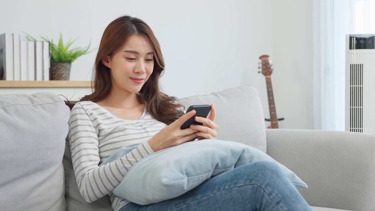 亚洲美女坐在沙发上，在家里用手机聊天。快乐迷人的年轻女孩在家里度过休闲时光，放松心情，享受在客厅用智能手机交流和讨论。视频素材