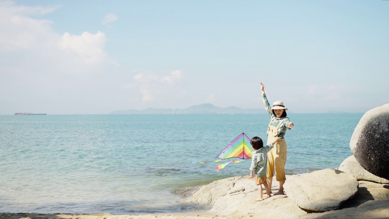 亚洲母亲和她的小儿子在休闲布在岩石海滩上玩彩色的风筝在夏天，有质量的时间在一起视频素材