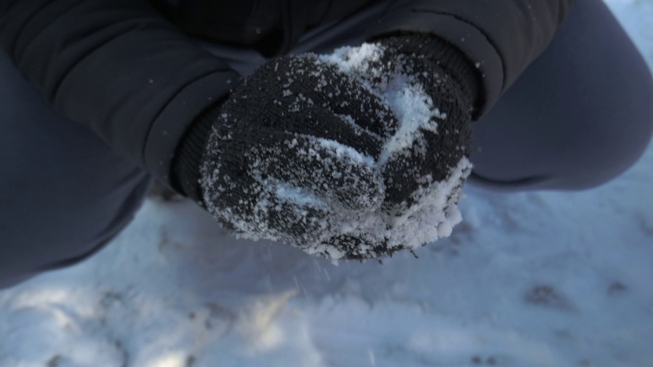 一个人的手在做雪球的特写镜头。这个人戴着黑色的羊毛手套，穿着黑色的夹克和蓝色的运动服视频素材