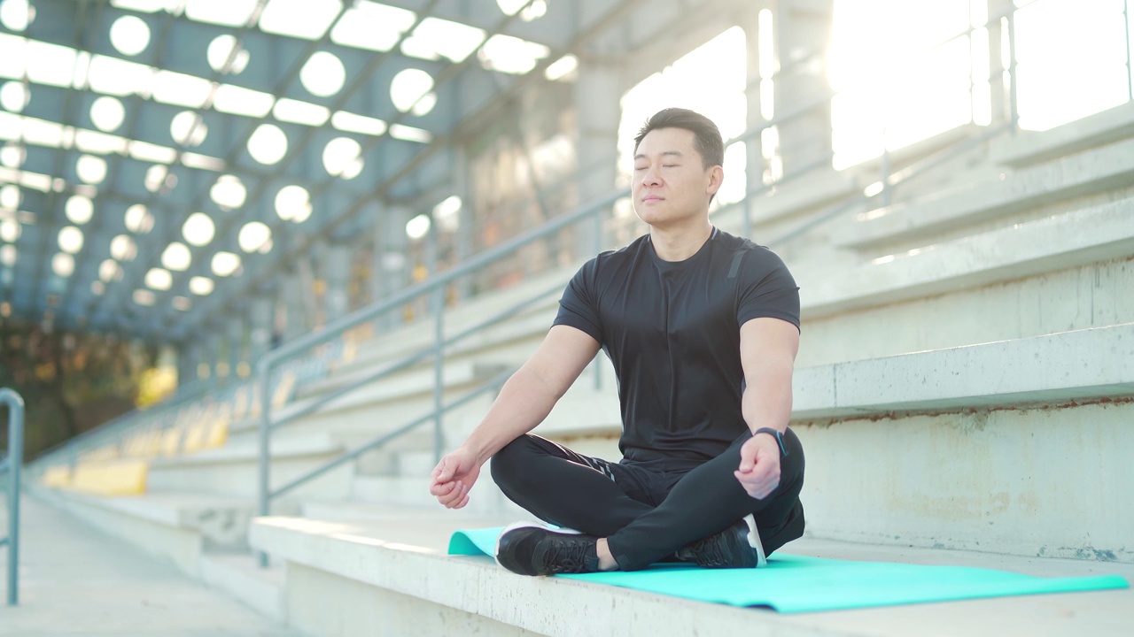 英俊的亚洲男子冥想在现代城市体育场，坐莲花姿势练习瑜伽垫，视频素材