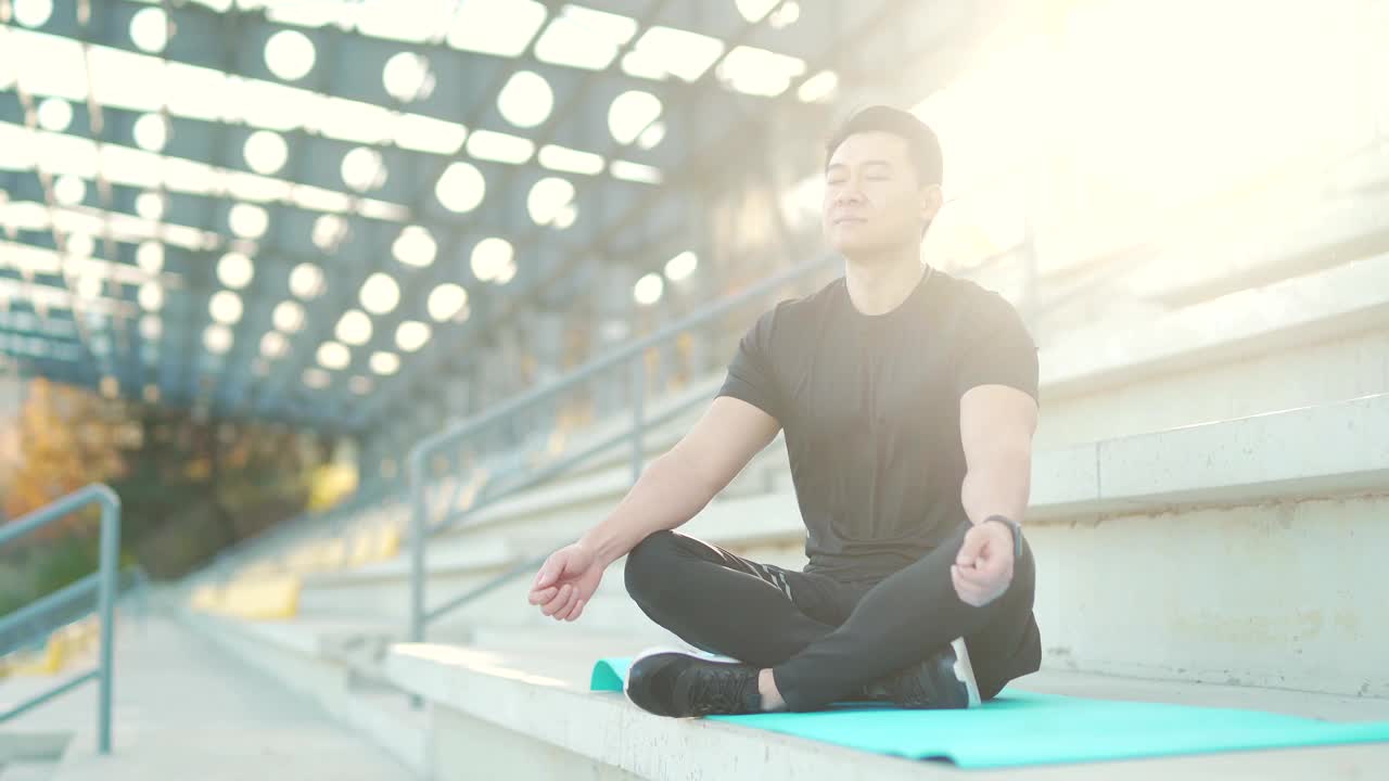 英俊的亚洲男子冥想在现代城市体育场，坐莲花姿势练习瑜伽垫，视频素材