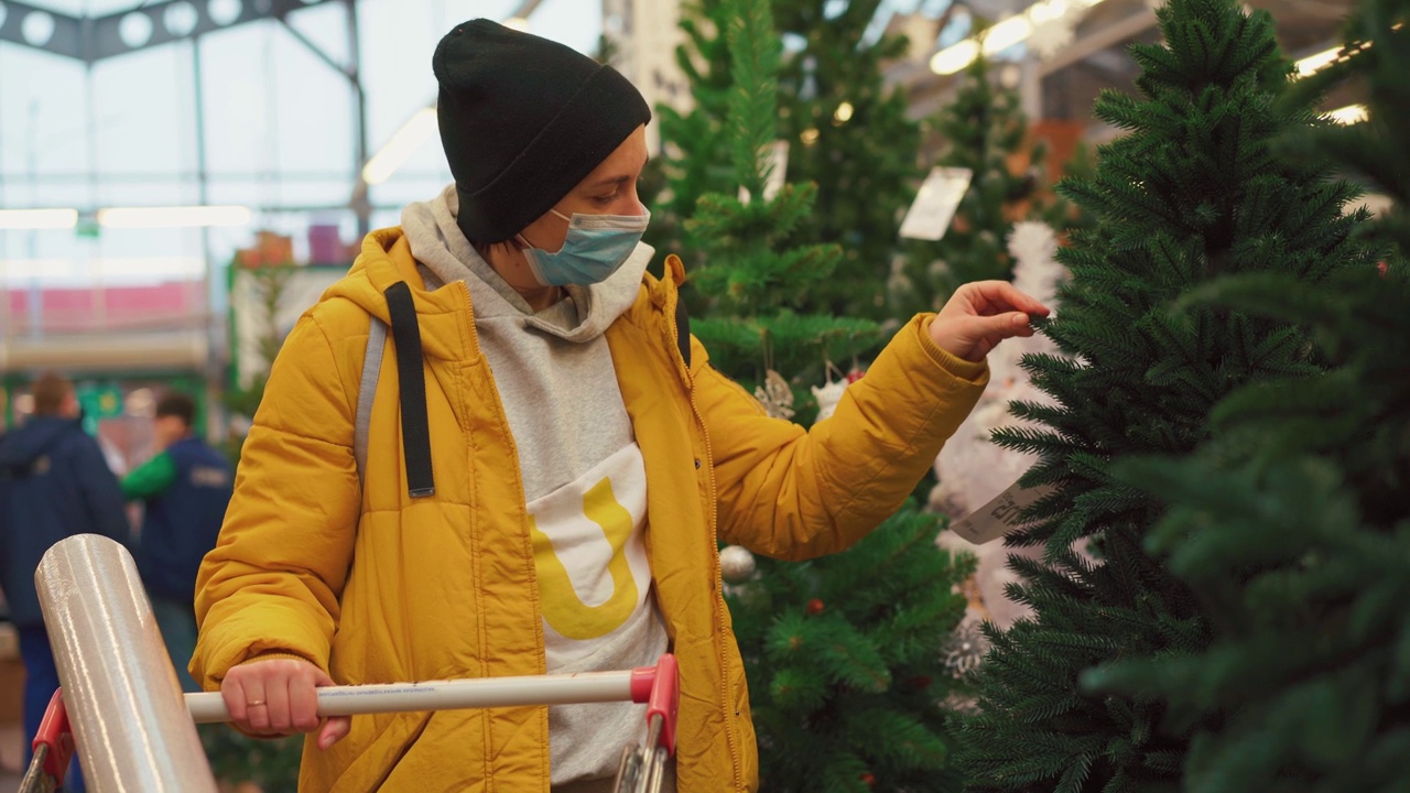 妇女在市场上挑选圣诞树。一名白人女性穿着黄色的冬季夹克，戴着医用防护口罩，在购物中心观看触摸人造圣诞树视频素材