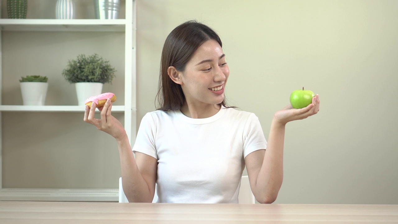 亚洲年轻女子节食。在甜甜圈和苹果之间选择。选择食物对你的健康有好处。健康的食物选择。视频素材