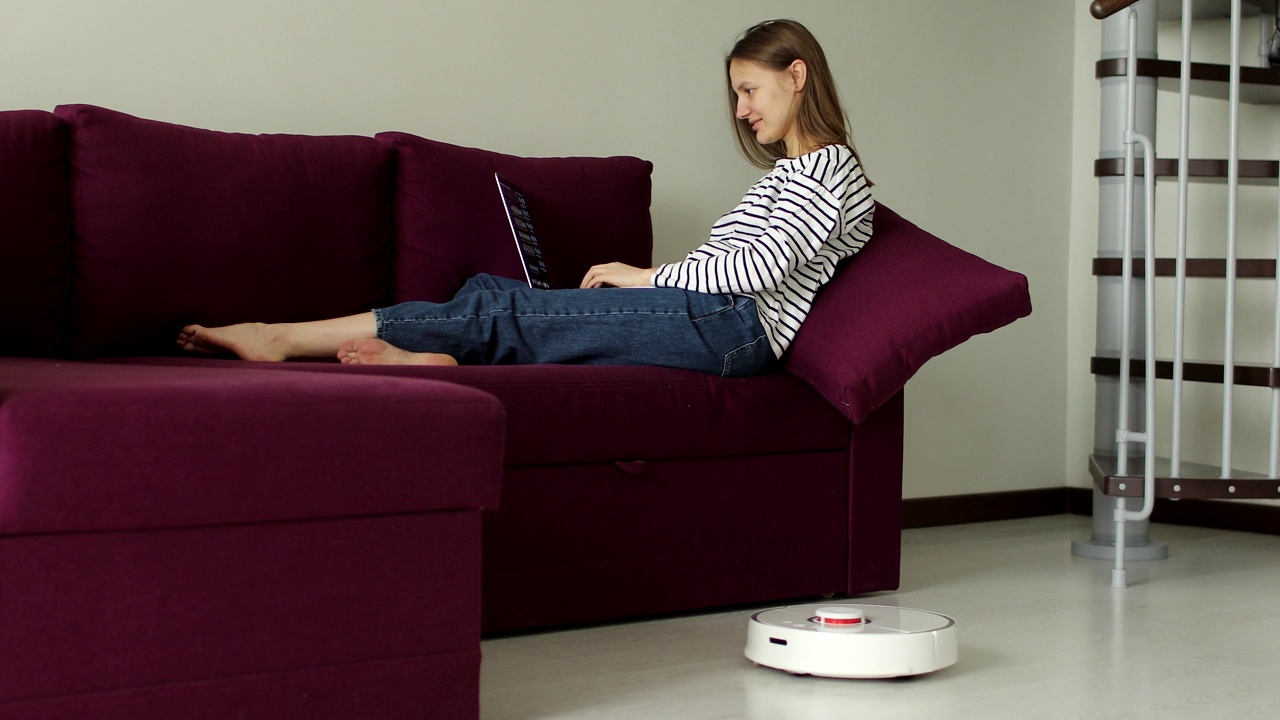 当女性在家庭办公室的沙发上工作时，智能吸尘器通过清洁地板。卫生、家居用品和机器人在现代生活中的概念。视频素材