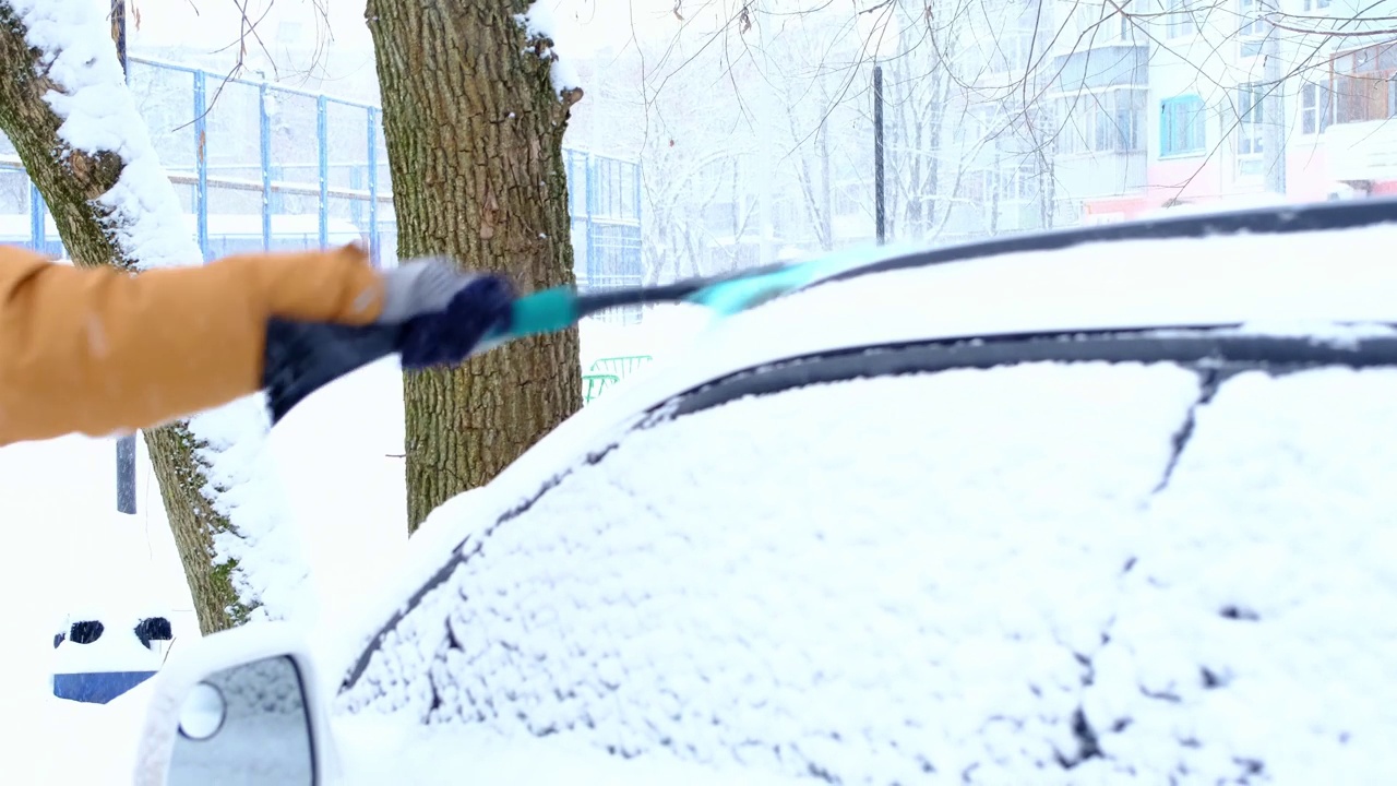 一名男子在雪后从车上刷雪。一只穿着芥末色夹克的手，白色的身体上放着汽车扫把。冬天的天气条件。慢动作视频素材