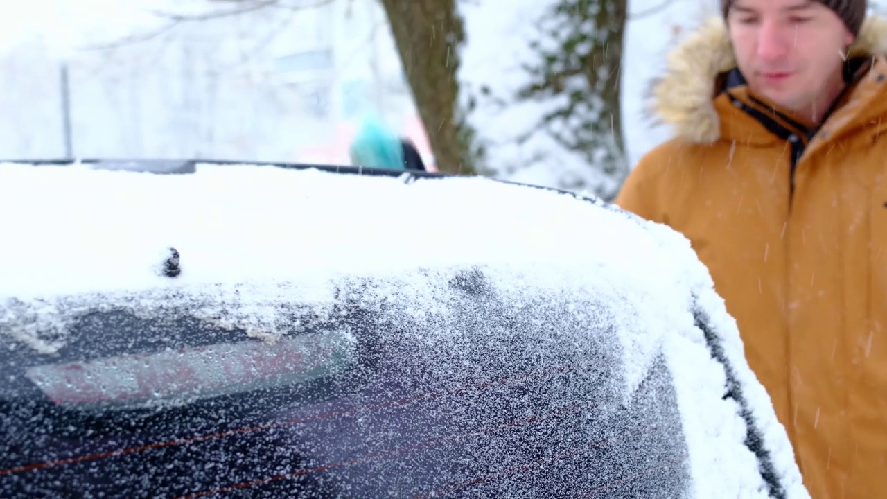 一名男子在雪后从车上刷雪。一只穿着芥末色夹克的手，白色的身体上放着汽车扫把。冬天的天气条件。慢动作视频素材