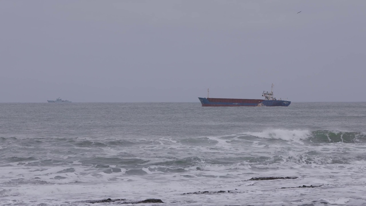 在暴风雨天气下波涛汹涌的海面上，一艘油轮沿着一艘领航船和一艘军用船的镜头在背景中出现视频下载