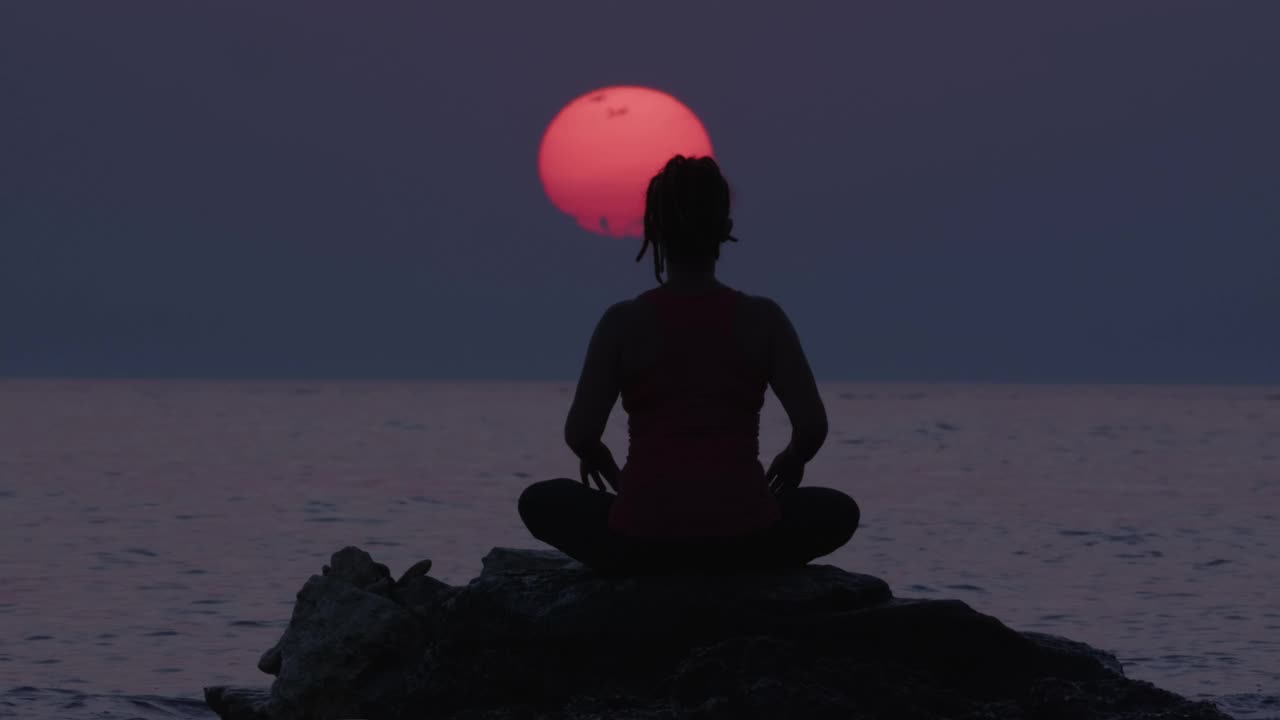 在令人惊叹的日落天空和大太阳期间，坐在海水中的石头上冥想的女人的剪影。治疗身体，心理和精神状态的人。视频素材