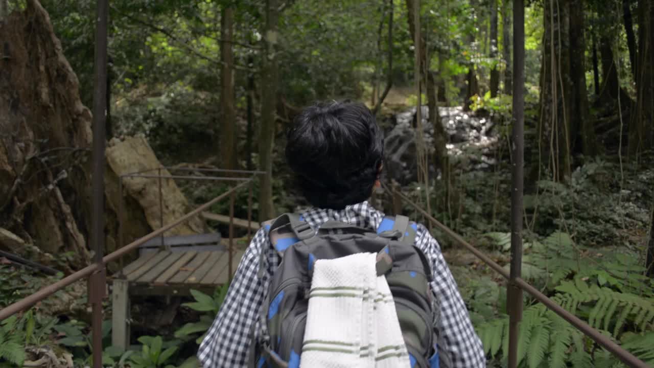 背后拍摄的女性自然旅行者背着背包走在木桥上，目的是在热带森林的瀑布。徒步穿越绿色植物和美丽的丛林。视频素材