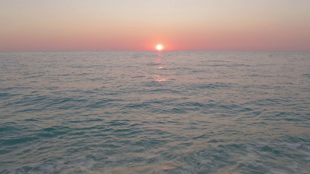 空中飞过那个坐在海滩上的男人的剪影。海水表面平静的波浪在日落黄昏发光的橙色天空。视频素材