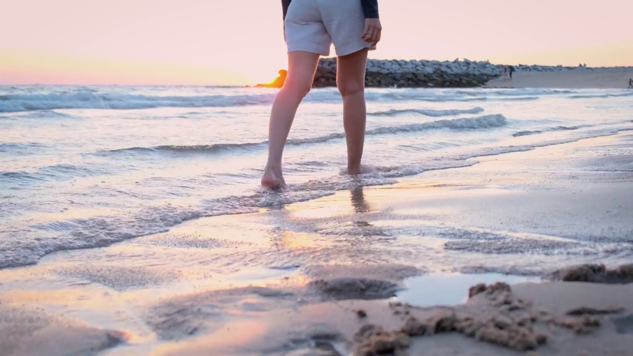 一个年轻的女人赤脚走在沙滩上，在沙滩上和柔软的海浪上留下脚印。在傍晚的时候，夕阳西下，天空的金色光芒给人一种温暖和平和的感觉。视频素材