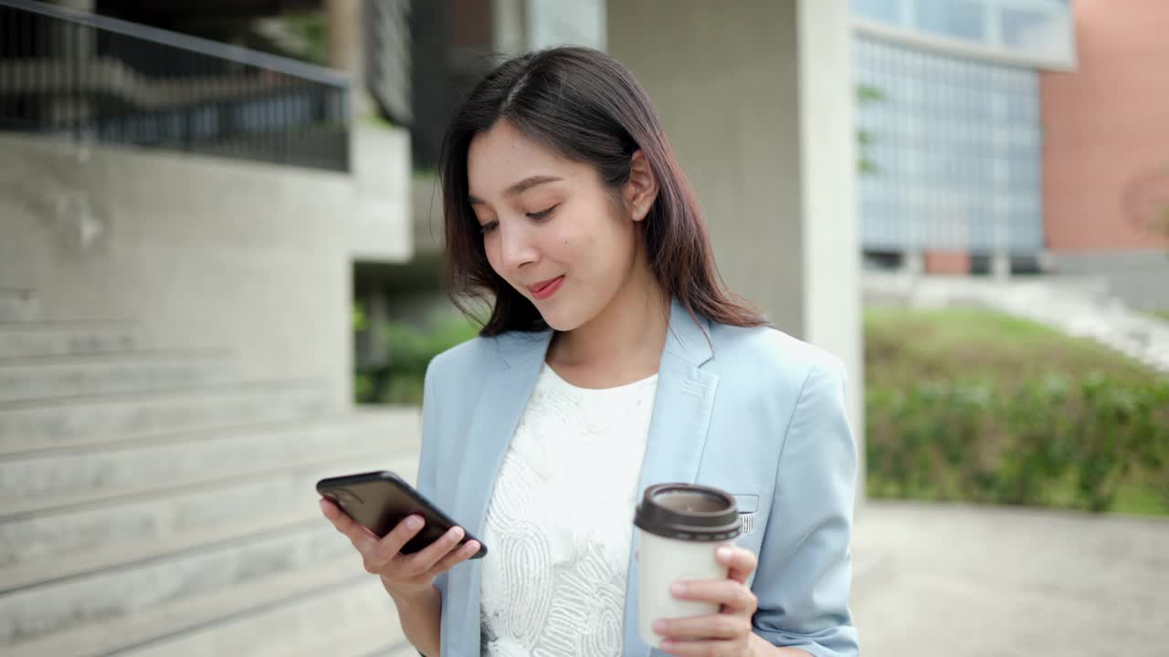 聪明的女性早上拿着智能手机和咖啡杯去办公室。漂亮的女商人拿着手机站在走廊之间的建筑物在市中心。视频素材