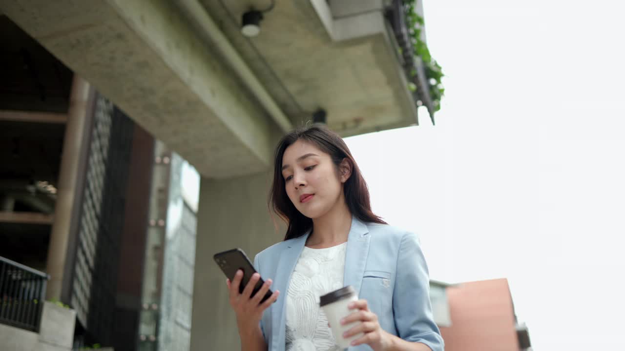 聪明的女性早上拿着智能手机和咖啡杯去办公室。美丽的女商人与移动电话行走在走廊之间的建筑物在市中心。视频素材