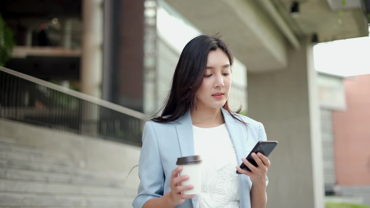 聪明的女性早上带着智能手机和咖啡杯去办公室。漂亮的女商人拿着手机站在走廊之间的建筑物在市中心。视频素材