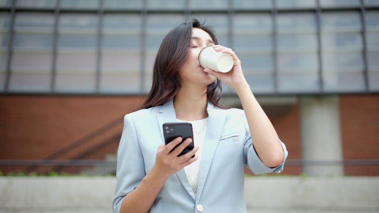 聪明的女性早上带着智能手机和咖啡杯去办公室。漂亮的女商人拿着手机站在走廊之间的建筑物在市中心。视频素材