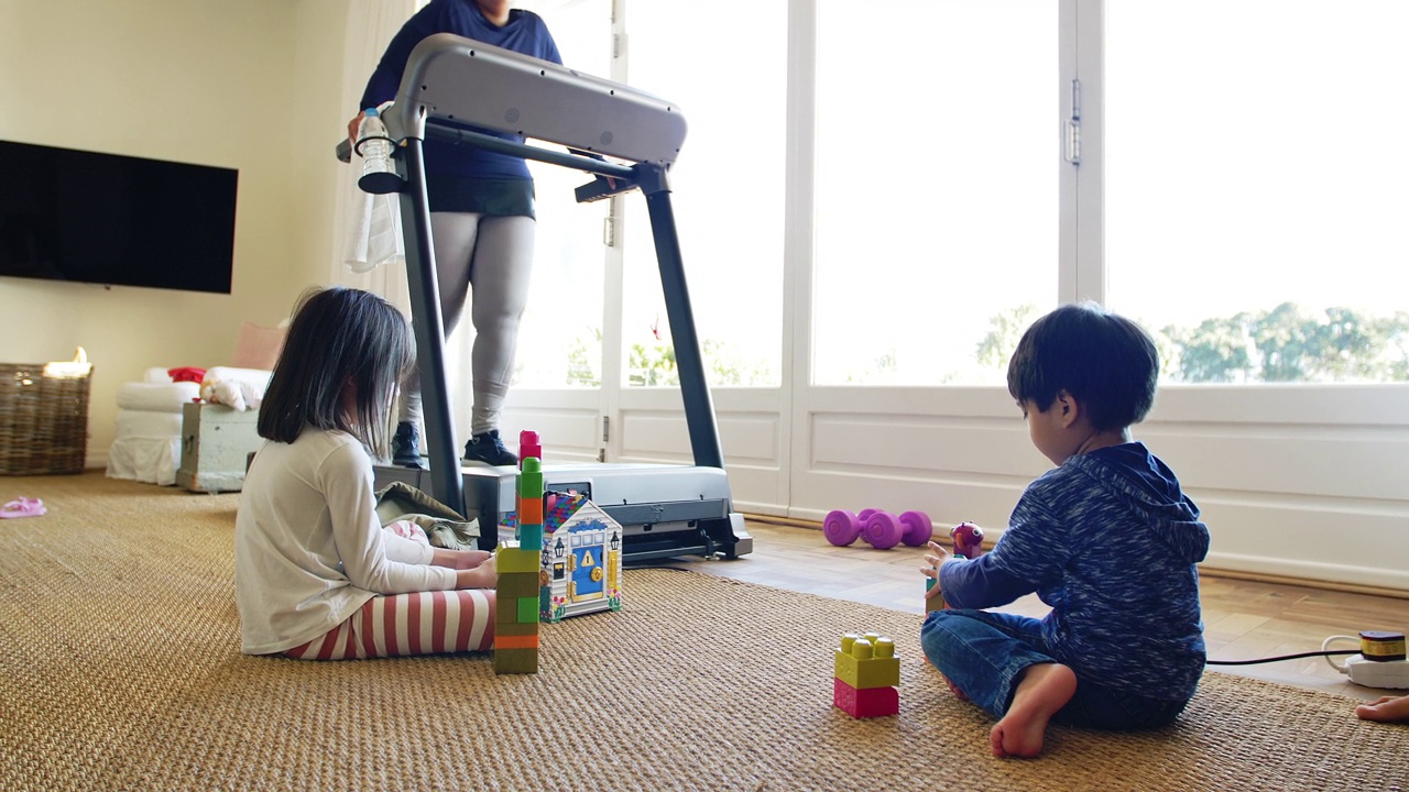 孩子们在地板上玩玩具，妈妈在跑步机上锻炼视频素材