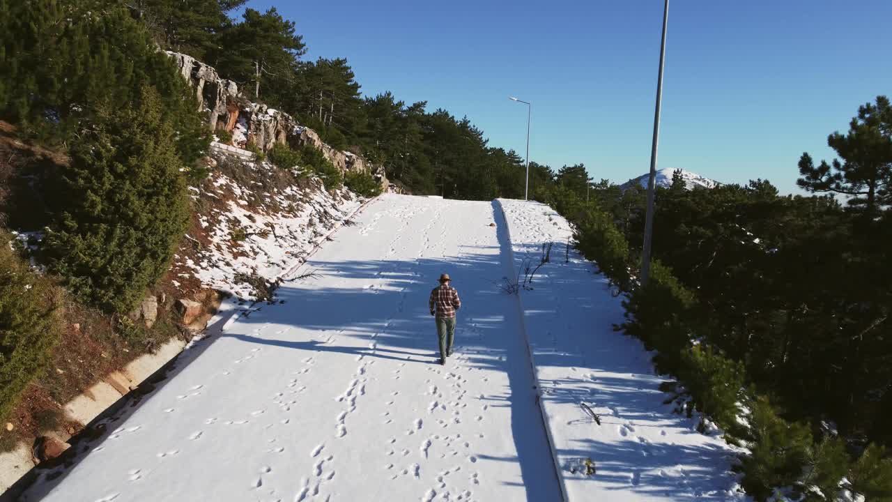 鸟瞰图，一个男人与软呢帽和棕色的伐木工人衬衫走在积雪的道路上的松树山。视频素材