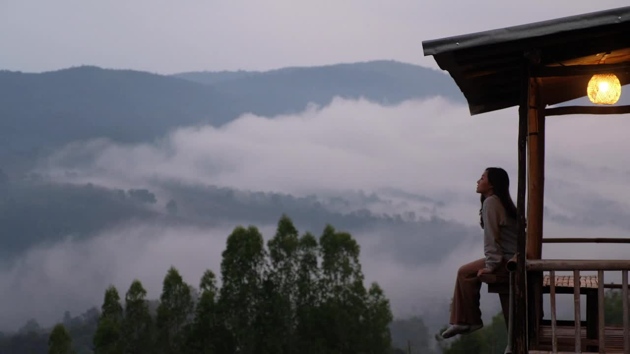 雾天，一位女旅行者坐在木屋阳台上，边喝酒边看美丽的山景视频素材