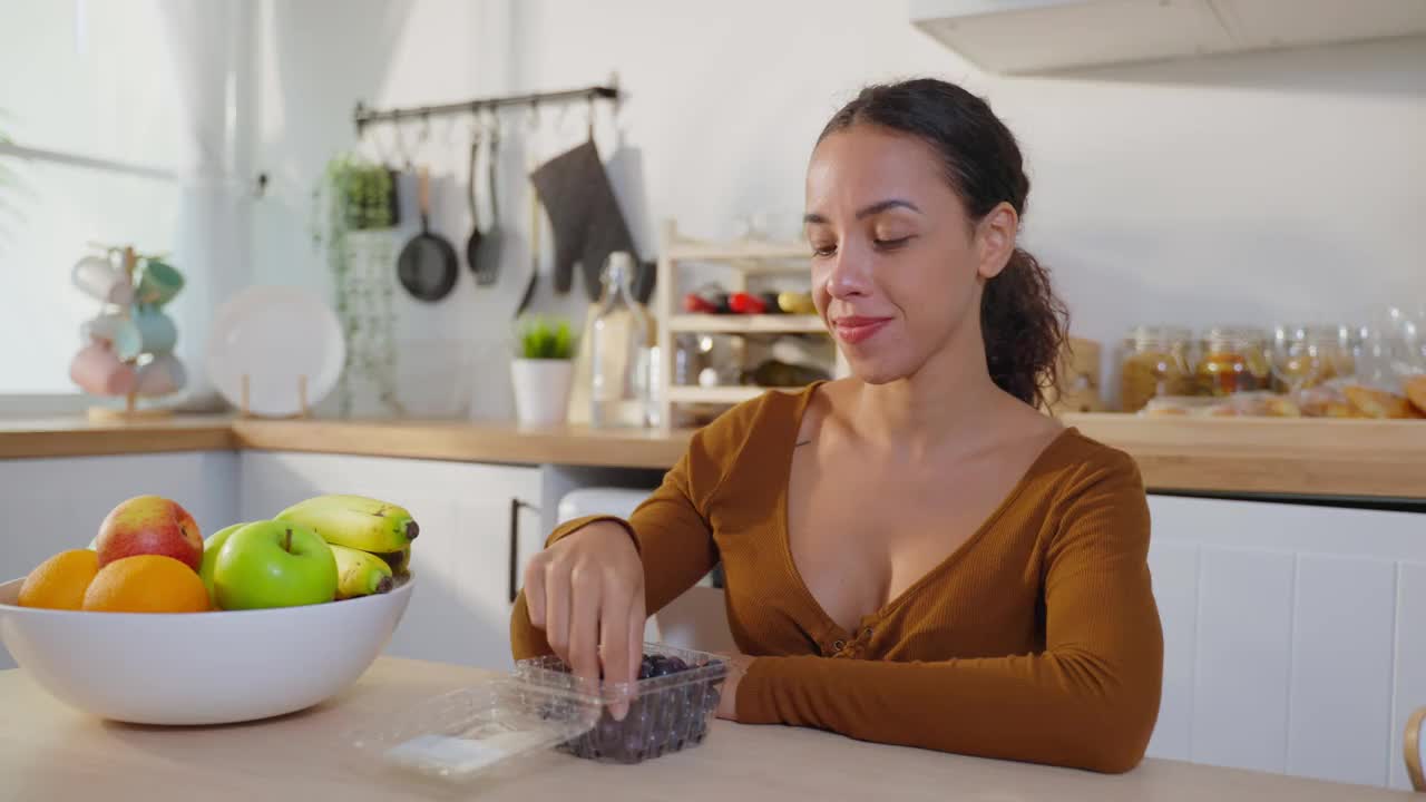 拉美美女喜欢在家里的厨房里吃健康的食物。美丽的年轻女性坐在餐桌上，吃着水果，喝着干净的水，在家保健减肥，感到很开心。视频素材