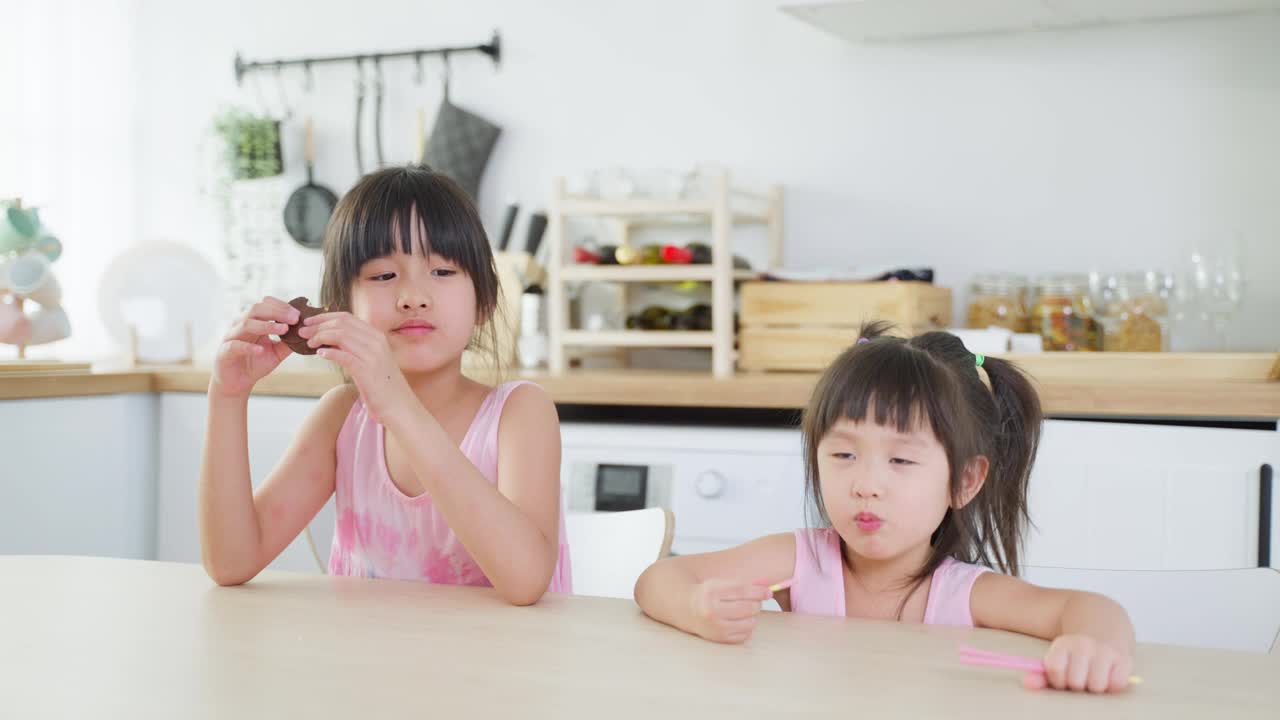 亚洲两个可爱的小女孩姐妹喜欢吃快餐，薯片。饥肠辘辘的学前班小女孩坐在厨房的桌子上，用手把零食放进嘴里。美味又不健康的儿童食品理念。视频素材
