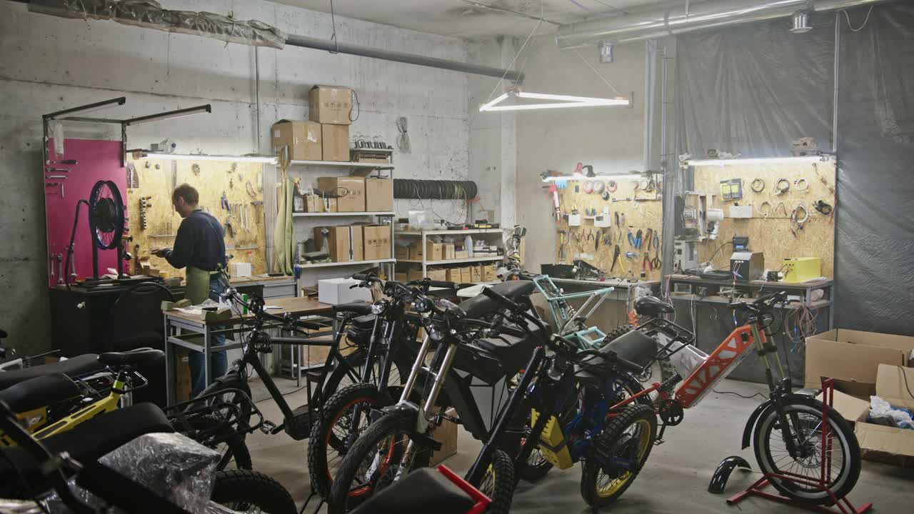 大型工场商业生态项目，用于生产电动自行车。一名男子在一个装有人工照明的大房间里，在一大堆电动自行车和环保自行车的背景下工作视频下载