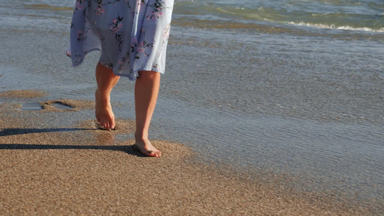 阳光明媚的夏日，沙滩上赤脚行走的女人留下了脚印。女孩在美丽的夏天摇摆着连衣裙走在海滩上日落视频素材