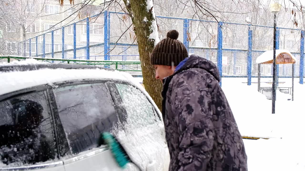 一名男子在雪后从车上刷雪。夹克里的一只手，白色的身体上放着一把汽车扫把。冬天的天气条件。慢动作视频素材