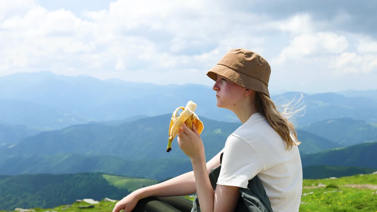 旅行者探险家女孩吃香蕉水果在一个阳光明媚的夏日的山顶上放松。成功、流浪和运动理念。视频素材